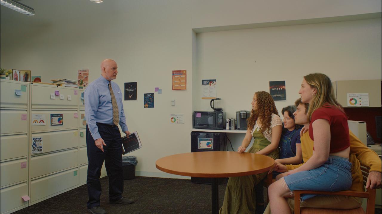 Craig standing over the four at the office table, delivering the news